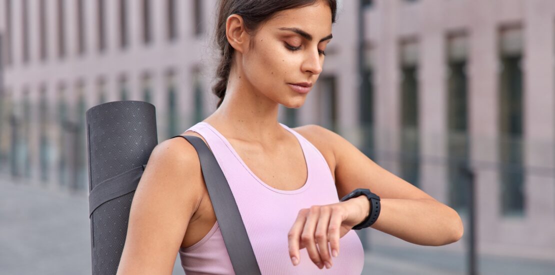 Outdoor shot of active young woman checks fitness results on fitness watch carries karemat dressed i
