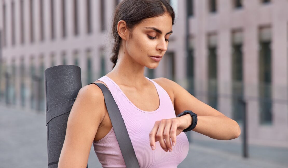 Outdoor shot of active young woman checks fitness results on fitness watch carries karemat dressed i