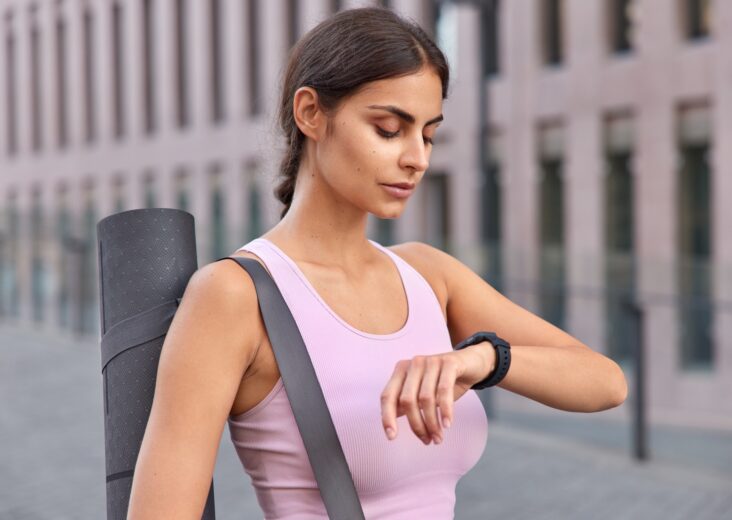 Outdoor shot of active young woman checks fitness results on fitness watch carries karemat dressed i