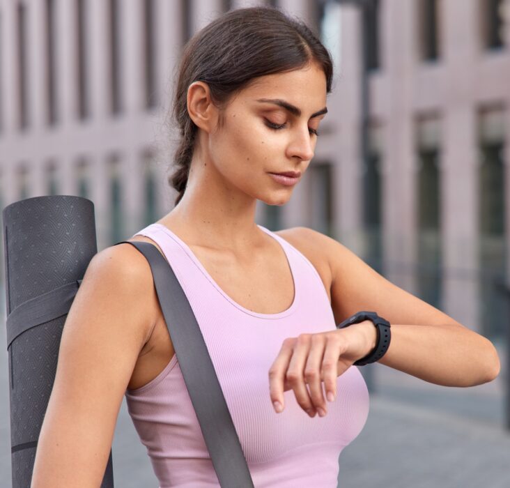 Outdoor shot of active young woman checks fitness results on fitness watch carries karemat dressed i