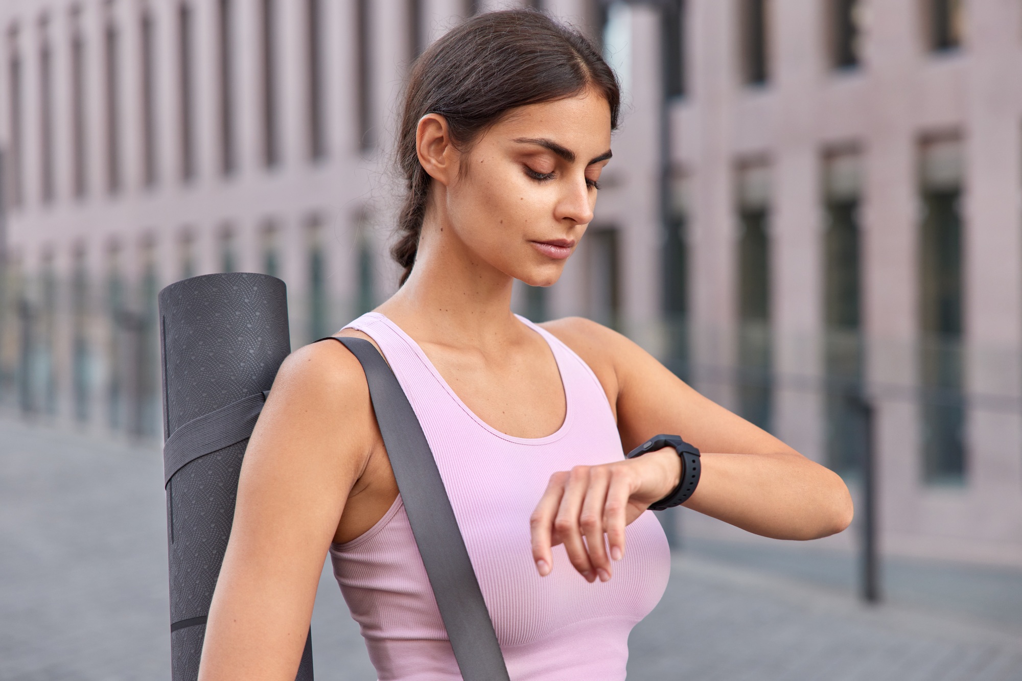 Outdoor shot of active young woman checks fitness results on fitness watch carries karemat dressed i